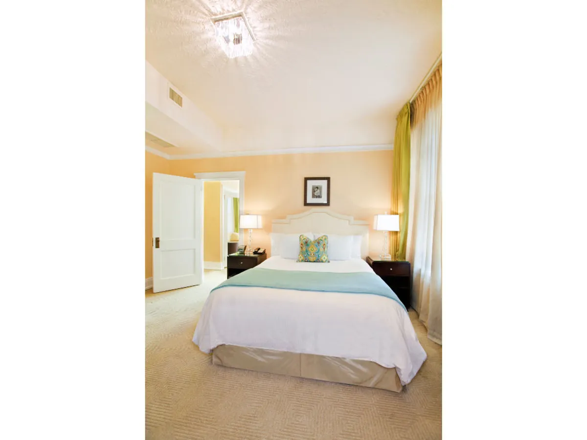A serene queen room scene with a welcoming bed dressed in premium bedding, complemented by nightstands, soft lighting from bedside lamps, and framed artwork above the headboard. Sunlight gently filters through curtain panels, creating an inviting, restful atmosphere.
