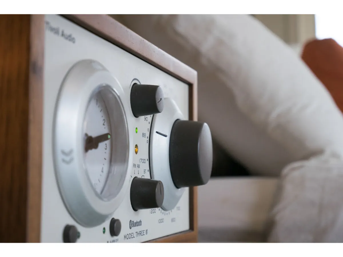A close-up of a vintage-inspired Bluetooth audio receiver resting on a wooden surface beside plush pillows, blending modern amenities with nostalgic charm. The soft, inviting textures and subtle design cues reference the era of classic Hollywood.
