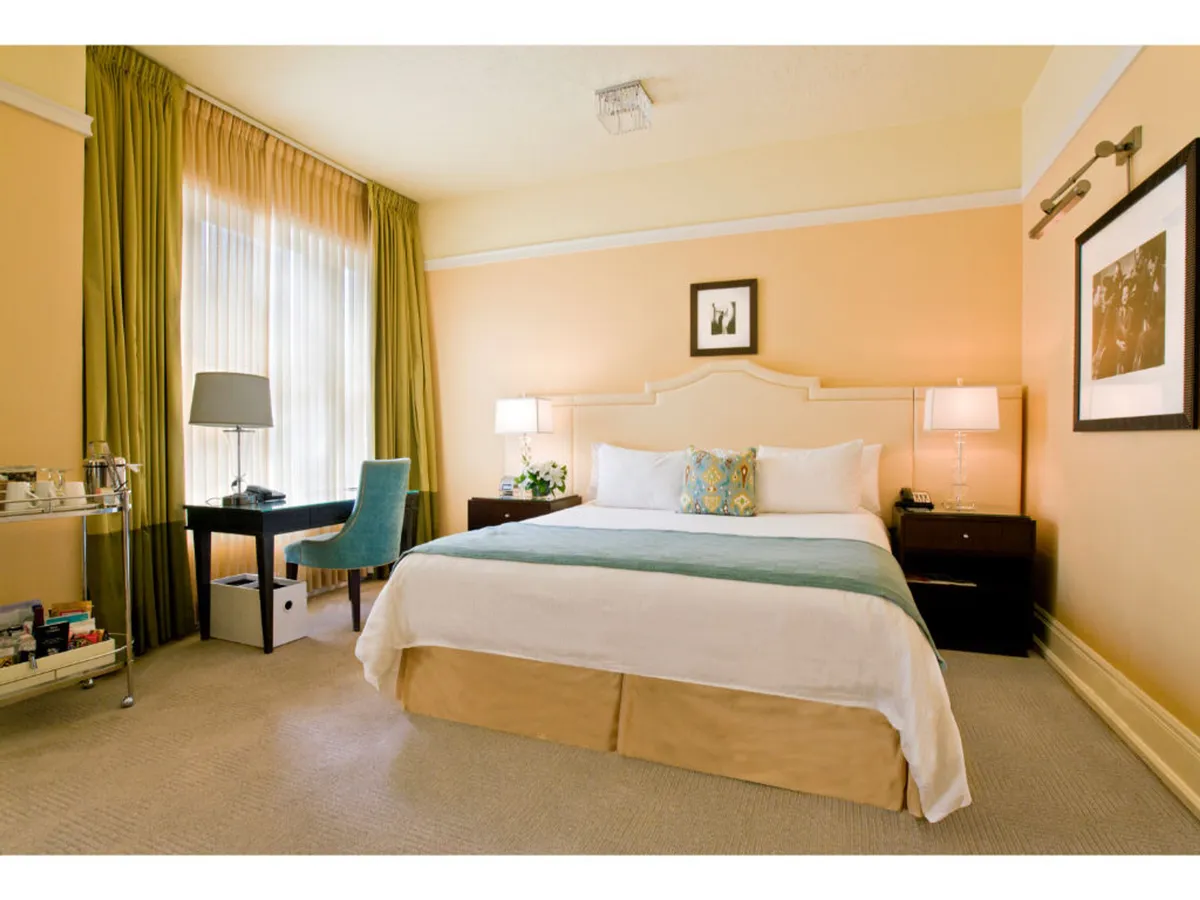 A sun-filled Premium Queen Room features a plush queen-sized bed adorned with decorative pillows, flanked by elegant side tables and lamps. Large windows with flowing drapes let in abundant natural light, while a sleek desk and chair provide a stylish workspace. Soft hues and understated details evoke an air of classic Hollywood glamour.