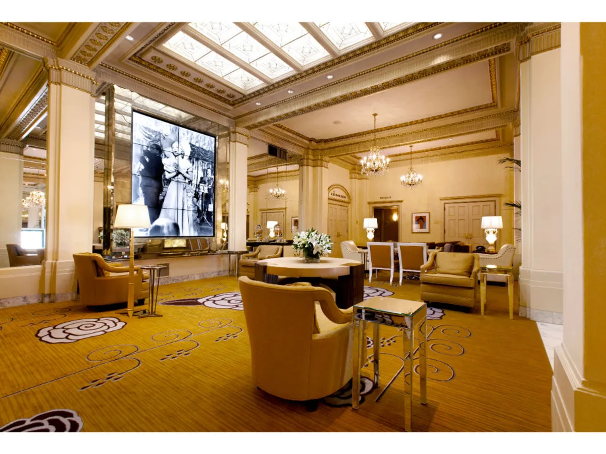 A stylish lounge area featuring a prominent large screen displaying a historical black and white image, a central round table with a floral arrangement, and inviting seating clusters. The ambiance is sophisticated yet welcoming, suggesting a communal space ideal for relaxation or socializing after a day’s adventure.