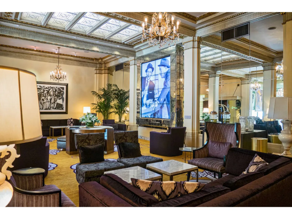 A charming, stylish interior highlighted by a central chandelier, large framed artwork, plush seating, tasteful greenery in large potted plants, and a screen displaying a historical image. The setting is ideal for both social gatherings and business meetings, with an aesthetic that feels sophisticated and welcoming.