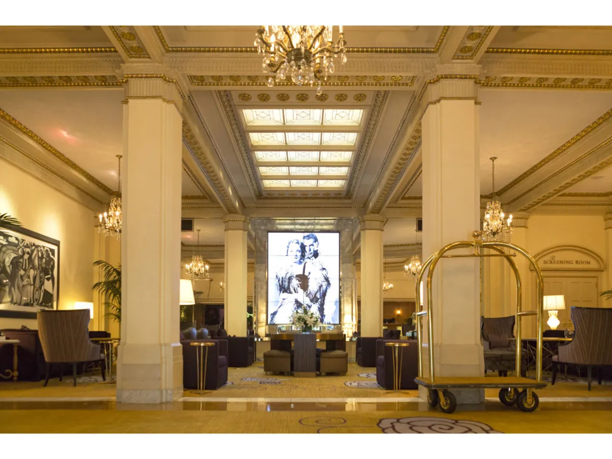 The interior scene highlights a luxurious lobby with grand chandeliers, a well-designed reception area, comfortable seating, large artwork, and a luggage cart ready for guests. The space feels both opulent and inviting, ideal for business meetings, casual relaxation, and artistic appreciation.