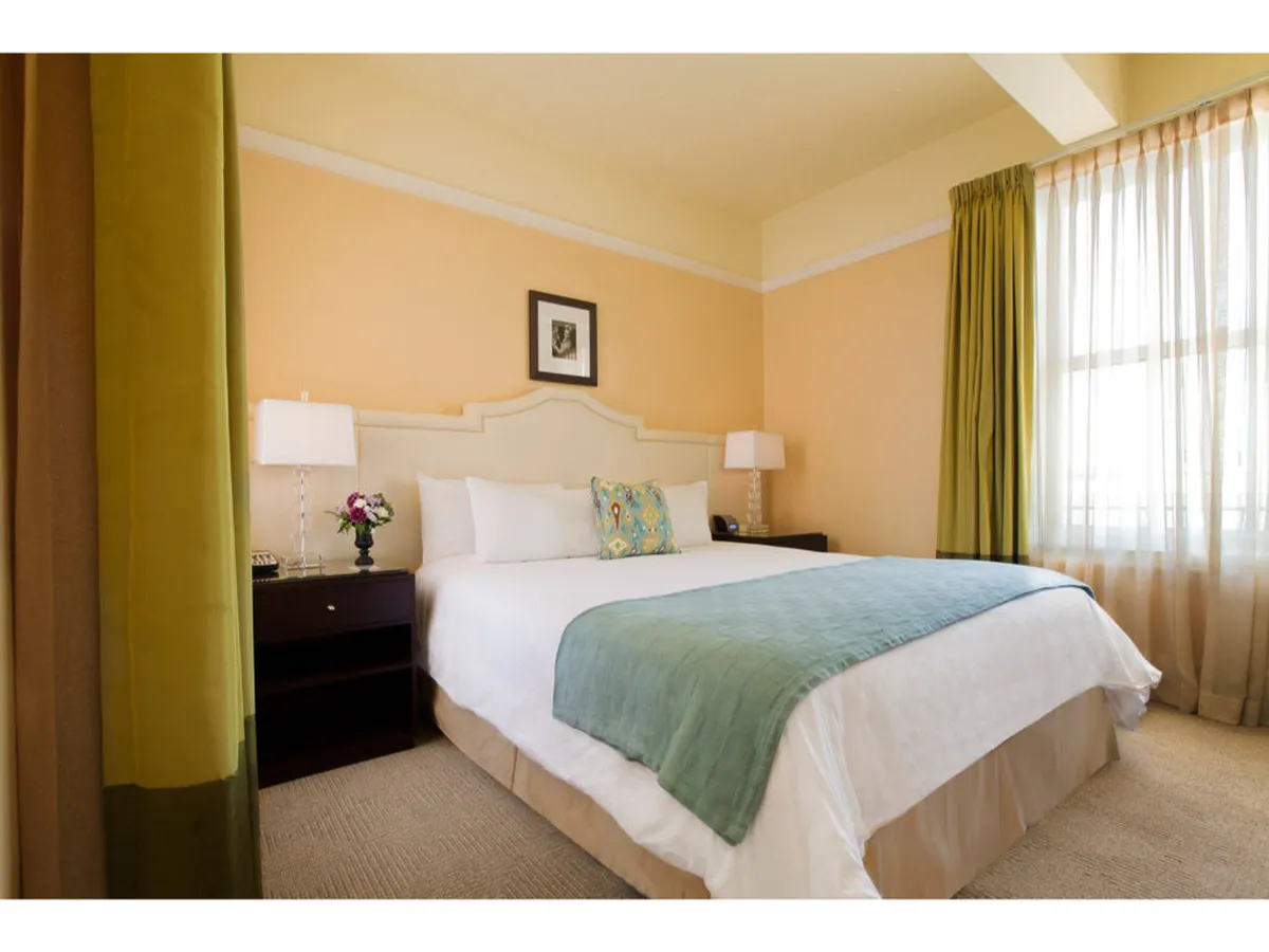 A beautifully appointed hotel room with a plush queen bed adorned with decorative pillows, elegant nightstands, and oversized window curtains gently filtering in soft morning light. The ambiance is calm and inviting, perfect for unwinding after a day exploring downtown Portland.