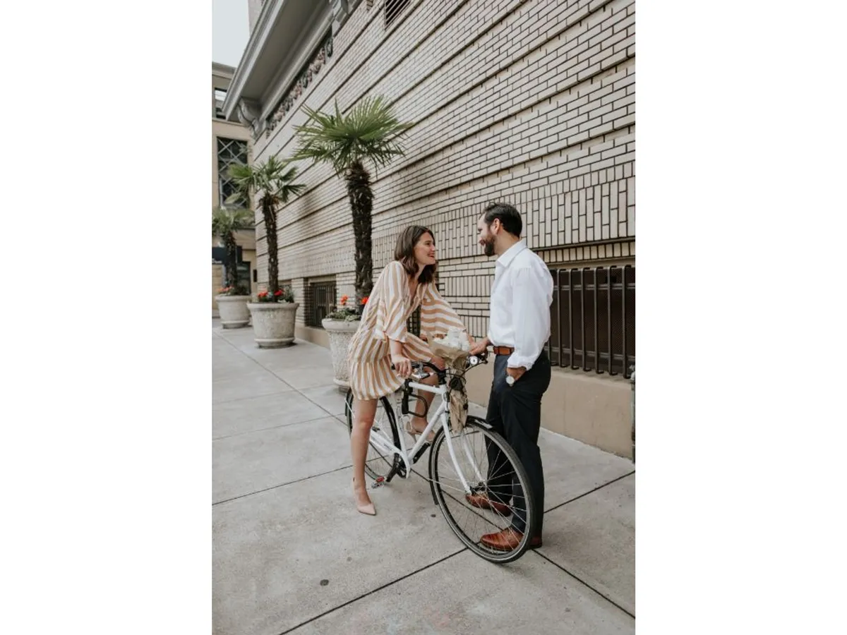 This exterior scene captures a couple interacting next to a bicycle outside the hotel, surrounded by potted plants. The atmosphere is playful and romantic, hinting at spontaneous urban exploration and active city adventures.